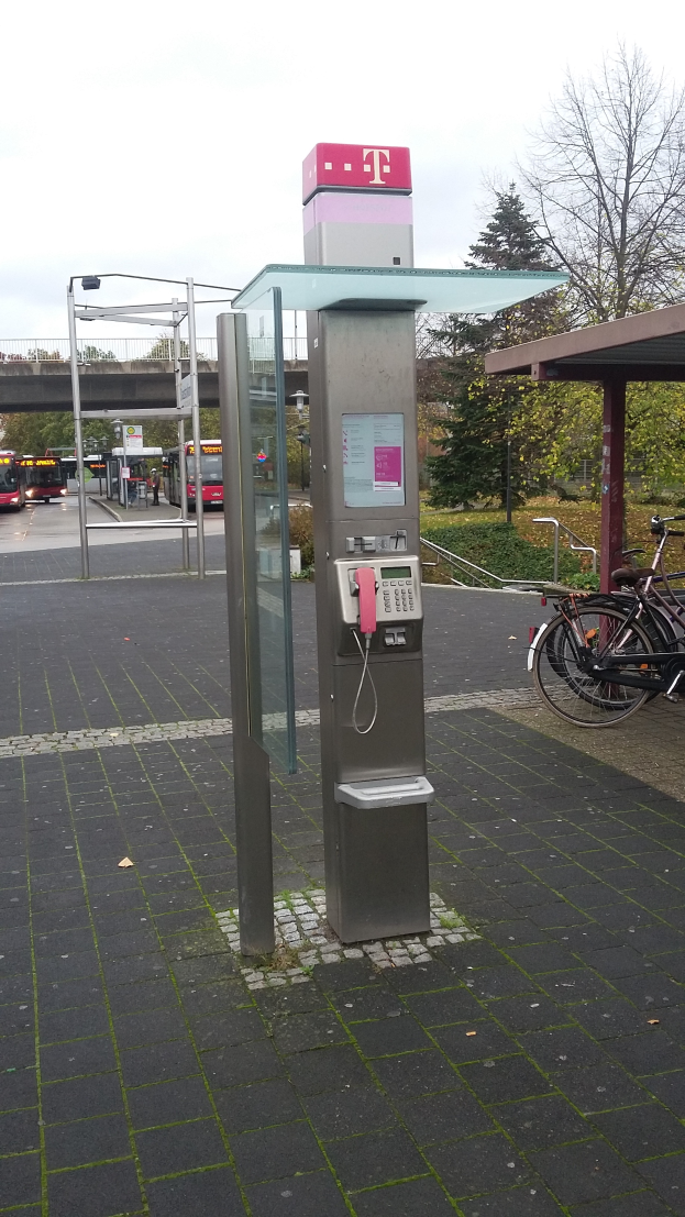 Ein Münzfernsprecher steht an der Seite einer Straße neben einer Bushaltestelle, daneben ist ein Fahrrad abgestellt; im Hintergrund gibt es Bäume, Fahrzeuge auf der Straße, eine Brücke und einen klaren blauen Himmel.