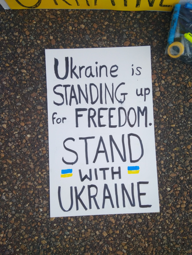 Weißer Zettel mit dem Text "Ukraine steht für Freiheit ein, steh mit Ukraine" auf dem Boden vor einer gelben Fahne, mit einer Plastikhülle mit verschiedenen Gegenständen rechts.