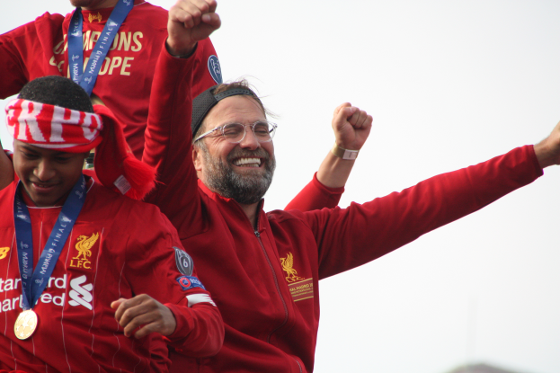 Liverpool-Manager Jürgen Klopp feiert mit seinen Spielern nach dem Gewinn der UEFA Champions League, während zwei medaillentragende Personen in der Nähe stehen und der Himmel im Hintergrund zu sehen ist.