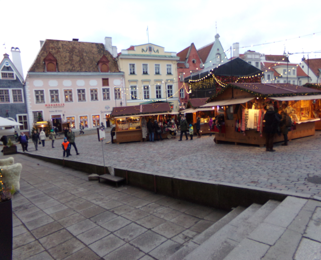 Ein geschäftiger Weihnachtsmarkt in Tallinn, Estland mit Menschen um geschmückte Stände, festliche Lichter, Gebäude im Hintergrund, bewölktem Himmel, Treppen nach oben und Topfpflanzen.