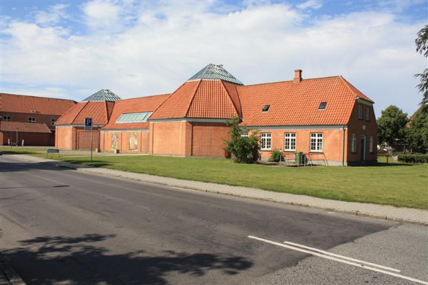 Ein großes Backsteingebäude mit rotem Dach, umgeben von Grünflächen und Schildern, das an der Seite einer Straße steht, unter einem bewölkten Himmel, repräsentiert die neue Schule in den Niederlanden.