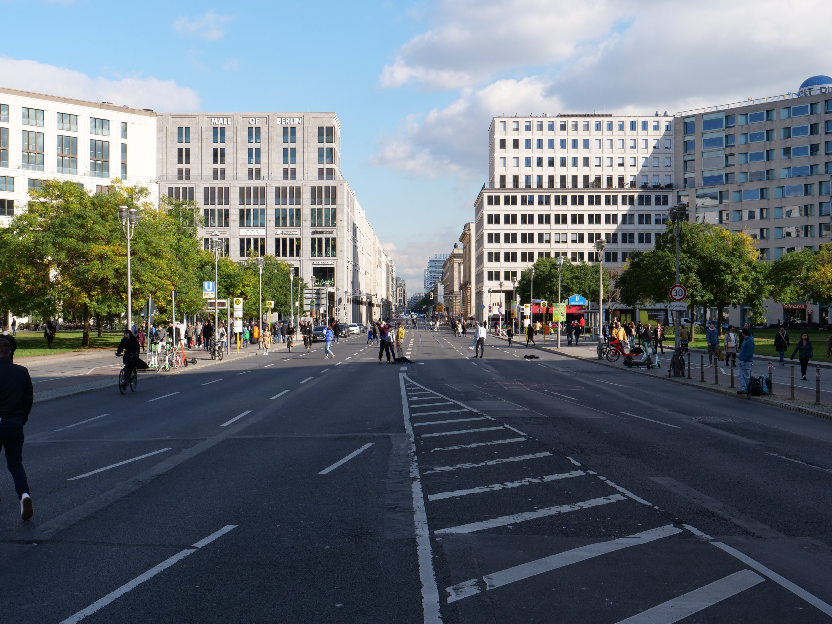 Eine belebte Straßenkreuzung in Berlin, Deutschland, mit Fußgängern und Radfahrern auf der Straße, hohen Gebäuden mit Fenstern, Bäumen, Laternen und Schildern, unter einem bewölkten Himmel.