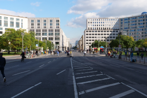 Eine belebte Straßenkreuzung in Berlin, Deutschland, mit Fußgängern und Radfahrern auf der Straße, hohen Gebäuden mit Fenstern, Bäumen, Laternen und Schildern, unter einem bewölkten Himmel.