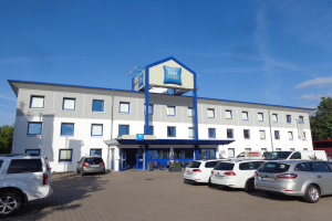 Ein Gebäude mit Fenstern und einem Schild mit der Aufschrift "ibis budget frankfurt airport", daneben geparkte Autos, Topfpflanzen, ein Straßenpfahl, Bäume und ein bewölkter Himmel.