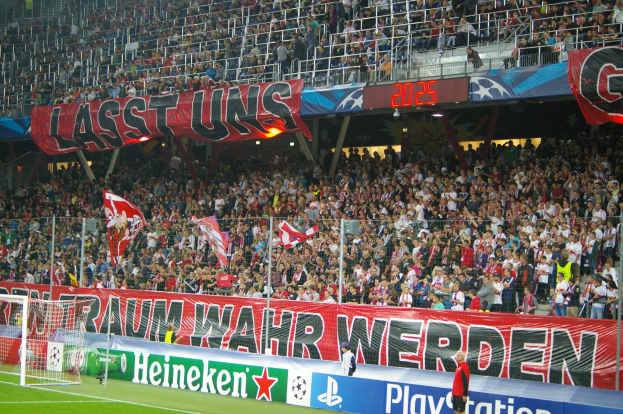 Ein Füßballspiel in einem Stadion mit einer großen Zuschauermenge, saftig grünem Rasen, einem Torpfosten links, Bodenbanner, Menschen mit Fahnen und Hintergrundgeländer, Tafeln, Lichter und andere Gegenstände.