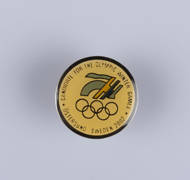 Ein rundes Abzeichen mit schwarzem Hintergrund und goldfarbenem Olympia-Logo, mit der Aufschrift "Olympic Winter Games" in fetter weißer Schrift.