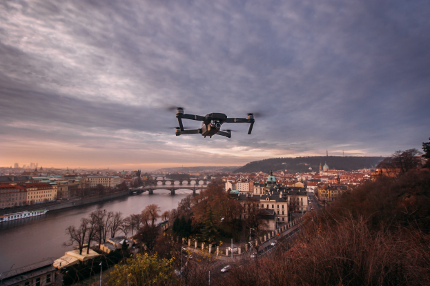 Ein Drohne fliegt über einer Stadt mit Gebäuden, Bäumen und Fahrzeugen, mit einem Fluss und einer Brücke im Hintergrund.