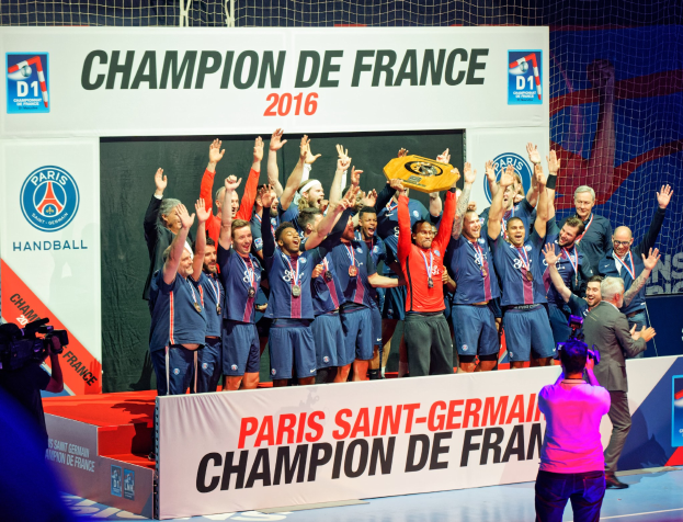 Eine Gruppe von Männern hält einen Pokal auf der Bühne, während eine Videokamera aufnimmt und ein 'Champion de France 2016'-Schild im Hintergrund zu sehen ist.
