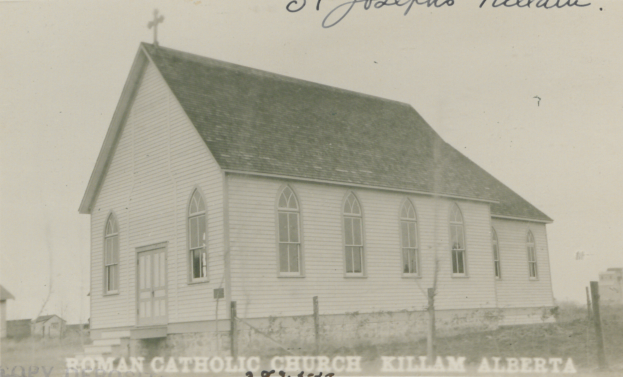 Ein Schwarz-Weiß-Foto von der lutherischen St. Joseph-Kirche in Killam, Alberta, das ein Haus mit Dach, Fenstern, Türen und einem Kreuz auf dem Dach zeigt, umgeben von einem Zaun, Gras und anderen Häusern im Hintergrund, mit Text oben und unten auf dem Bild.