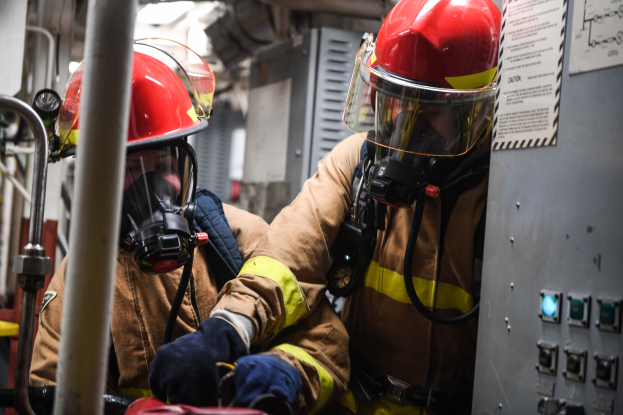 Zwei Feuerwehrmänner in Schutzausrüstung, einschließlich Helmen und Gasmasken, arbeiten an einem Feuerwehrauto, wobei eine Tafel mit Text und Metallstangen im Hintergrund sichtbar ist.
