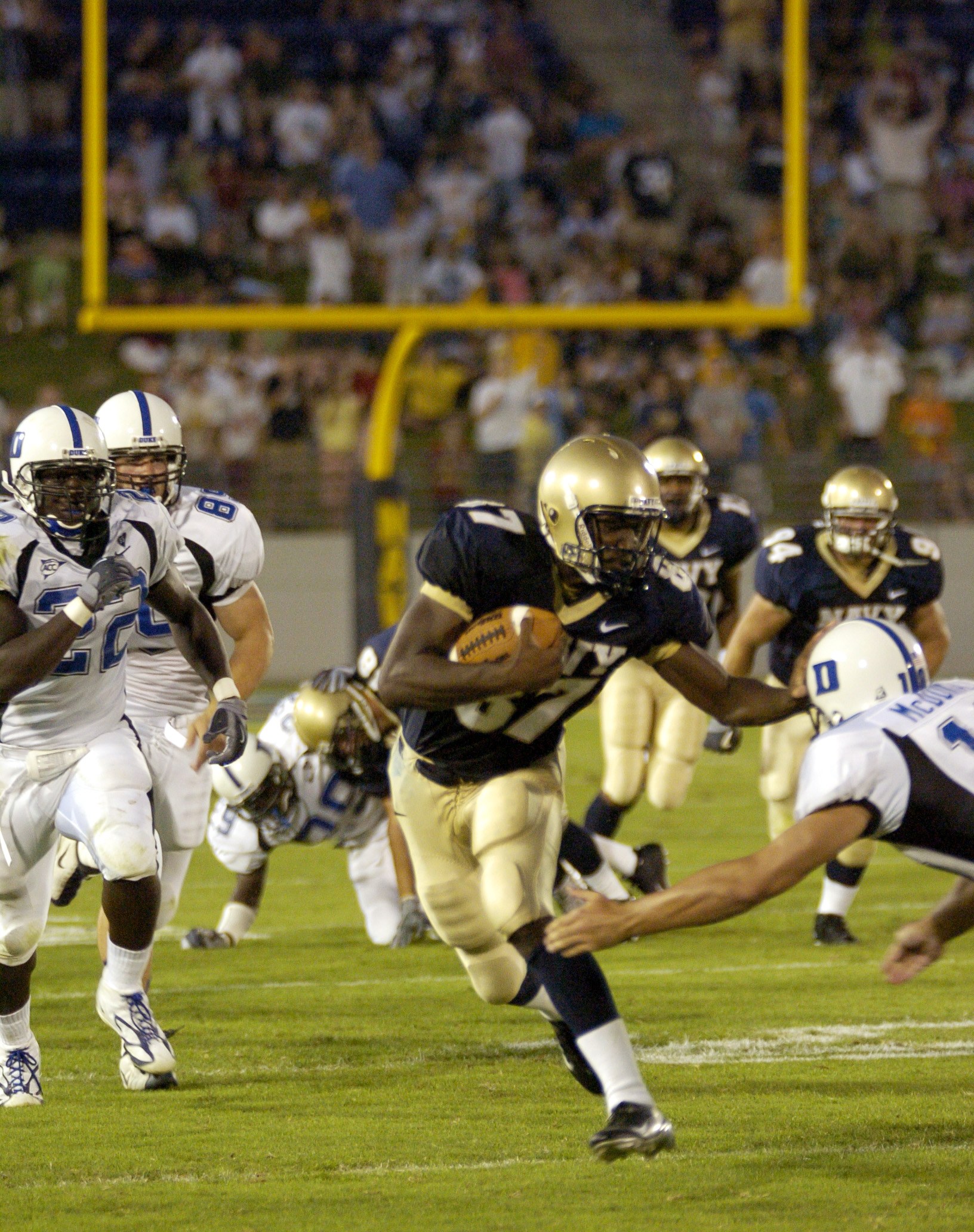 Fußballspieler in rotem Trikot mit Ball, verfolgt von Gegenspielern in Sportbekleidung und Helmen, mit unscharfen Zuschauern und Stadionelementen im Hintergrund und Text unten.