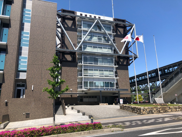Das Hauptquartier des Japanischen Olympischen Komitees, ein großes Gebäude mit zahlreichen Fenstern, ist von blühenden Pflanzen, Bäumen, Fahnenmasten und einem Fahrzeug auf der Straße umgeben, mit einer Brücke und einem klaren blauen Himmel im Hintergrund.
