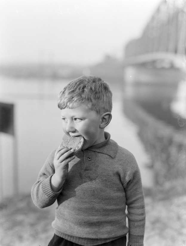 Schwarzes und weißes Foto eines jungen Jungen, der ein Stück Brot in der Hand hält und steht, mit einem unscharfen Hintergrund.