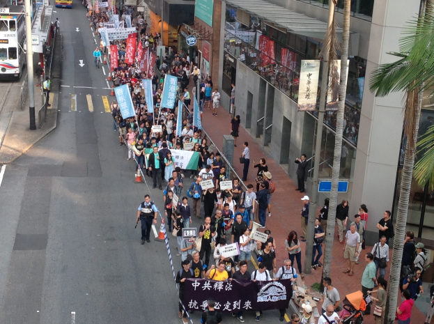 Große Gruppe von Menschen marschiert bei einer Demonstration durch eine Stadtstraße, hält Schilder und Plakate, mit Bäumen, Glasgebäuden und Fahrzeugen auf den Seiten.