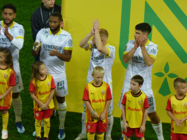 Eine Gruppe junger Menschen in Fußballtrikots steht auf einem Fußballfeld und klatscht feiernd mit einer 'Ligue 1'-Tafel im Hintergrund.