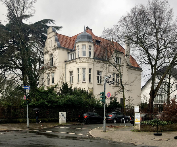 Ein großes weißes Gebäude mit einem roten Dach an einer Straßenecke, umgeben von Bäumen, Pflanzen und einem Zaun, mit Fahrzeugen, Verkehrszeichen und Schildern im Vordergrund und einem bewölkten Himmel im Hintergrund, das ehemalige deutsche Polizeipräsidium in Berlin.