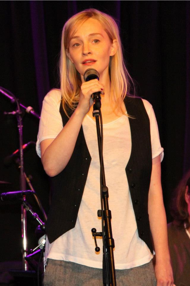 Frau auf einer Bühne mit Mikrofon stehend, die ein weißes T-Shirt und eine schwarze Jacke trägt, neben einer anderen Person und Musikinstrumenten im Hintergrund.