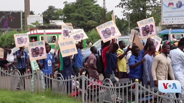 Eine Gruppe von Menschen hält Schilder in Händen vor einem Zaun, umgeben von Fahrzeugen, Bäumen, Masten, Werbetafeln und einem Bildschirm, mit Gras unten und dem Himmel im Hintergrund, scheinbar protestieren sie gegen eine Regierungentscheidung.