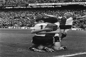 Drei Fußballspieler umarmen sich auf dem Boden beim Feiern eines Tors, mit jubelnder Menge und schwenkenden Fahnen im Hintergrund, dargestellt in Schwarz-Weiß.