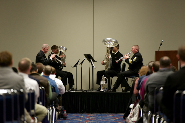 Eine Gruppe von Männern, die auf einer Bühne Blasinstrumente spielen, mit Notenständern, einem Podium mit Mikrofon und einer Sitzreihe im Publikum.