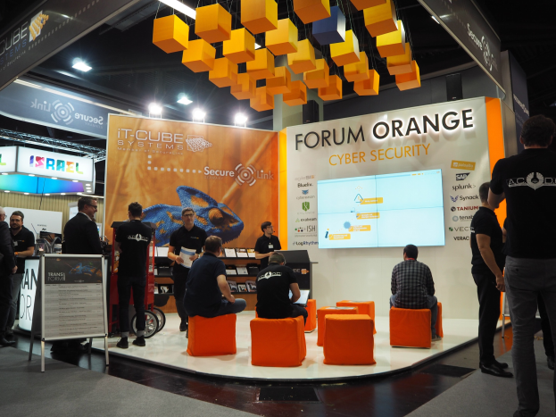 Eine Gruppe von Menschen an einem IT-Cube Systems Forum Orange Cyber Security Stand, mit sitzenden und stehenden Teilnehmern, einem Display-Bildschirm, Texttafeln, Regalen mit Gegenständen und Deckenleuchten.