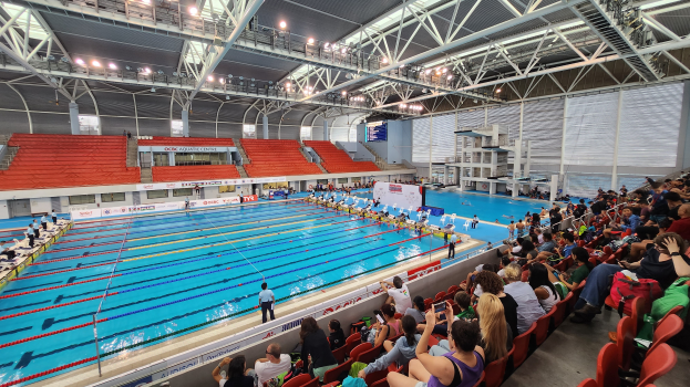 Großer Hallenbad mit vielen Menschen auf Stühlen drumherum, einige im Wasser stehend und Schilder mit Text im Hintergrund unter einer Decke mit Lampen und Eisenstangen.