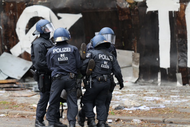 Polizeibeamte in schwarzen Uniformen und blauen Helmen stehen vor einem mit Graffiti bedeckten Gebäude während einer Unruhen, mit verstreuten Gegenständen und Text auf der Hintergrundwand.