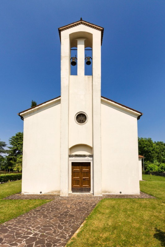 Kleine weiße Kirche mit einem zentralen Glockenturm, umgeben von einem Weg, Gras, Pflanzen, Bäumen und einem bewölkten Himmel, mit der laut klingenden Glocke.
