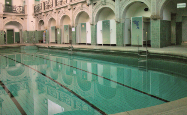 Innenbad mit kristallklarem Wasser, umgeben von einem gefliesten Boden, in einem großen Gebäude mit Bogengängen, Säulen, Geländern, Türen und Tafeln mit Text.