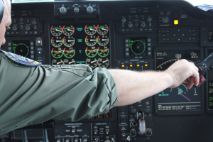 Ein Mann in einer Pilotenuniform sitzt in einem Flugsimulator-Cockpit und hält ein Objekt in der Hand, umgeben von verschiedenen Messgeräten und Bedienelementen.