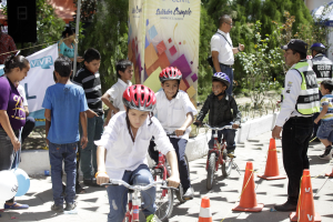 Kinder fahren mit Fahrrädern auf einer Straße mit Verkehrskegeln, einige tragen Helme, andere stehen in der Nähe, mit einem Banner, Bäumen und Gebäuden im Hintergrund.