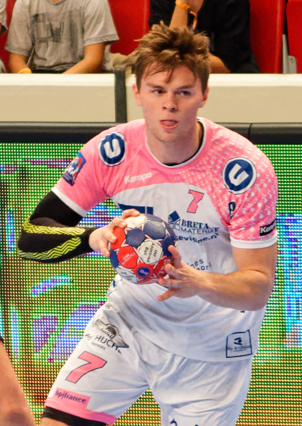 Ein Mann in einem rosa Hemd mit einem Fußball, mit der Hand eines anderen Menschen links daneben, während im Hintergrund Menschen auf Stühlen sitzen.