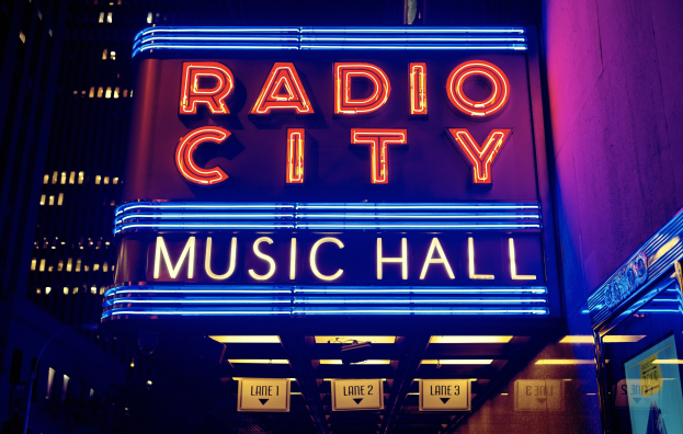 Außensicht des Radio City Music Hall in New York City bei Nacht, hell erleuchtet mit hohen Gebäuden im Hintergrund, mit einer "Radio City"-Leuchtschrift und mehreren kleineren Schildern auf der rechten Seite.