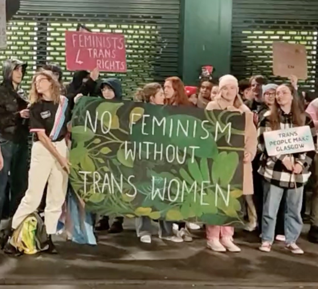 Eine Gruppe von Menschen mit einem Banner "Kein Feminismus ohne Transfrauen" und verschiedenen Schildern in einem öffentlichen Raum mit einer Wand und einem Rollladen im Hintergrund.