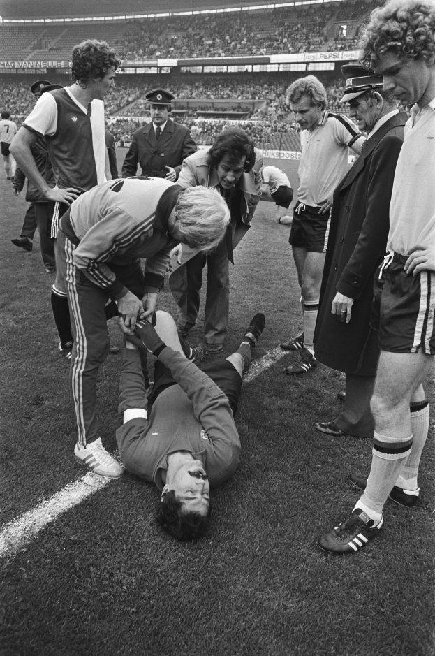 Schwarzes und weißes Bild eines Fußballspielers, der auf dem Boden liegt und von einer Gruppe Männer umgeben ist, mit einer Menschenmenge im Hintergrund.