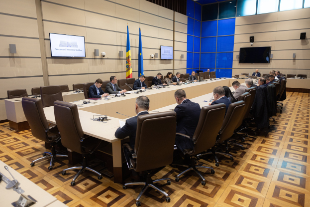 Große Gruppe von Menschen an einem langen Tisch mit Mikrofonen und Papieren in einer formellen Besprechungssituation mit Flaggen der Europäischen Union, Fernsehern, Deckenleuchten und Lautsprechern im Hintergrund.
