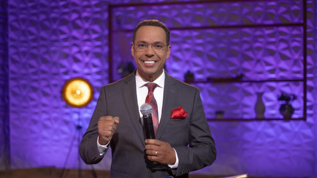 Stephen Colbert, Moderator von *The Daily Show with Trevor Noah*, steht auf einer Bühne und hält ein Mikrofon in der Hand, trägt einen Anzug, Krawatte und eine Brille, mit einer Wandmontage und einem Licht dahinter.