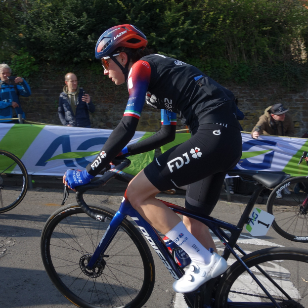 Eine professionelle Radsportlerin, Katie Archibald, fährt in Helm, Brille, Handschuhen und Schuhen ein Fahrrad eine Straße entlang neben einer Menschenmenge, mit einem Banner, einer Wand, Bäumen und einem Gebäude im Hintergrund.