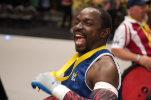 Ein Mann im Rollstuhl mit ausgestreckten Armen und einer Medaille um den Hals lächelt im Vordergrund, während im Hintergrund Menschen, Stühle und andere Gegenstände zu sehen sind.