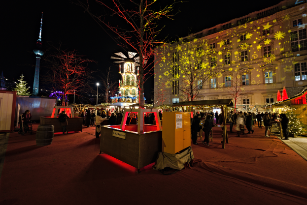 Ein lebendiger Weihnachtsmarkt in Berlin, Deutschland, mit Menschen um beleuchtete Stände, Bäume, Gebäude, Laternenmäste und einen Turm unter einem dunklen Himmel.