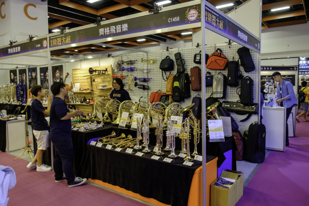 Gruppe von Menschen um einen Tisch mit Musikinstrumenten, Taschen und Schildern mit Text bei dem Hong Kong International Music Festival, mit Deckenbeleuchtung und einem Karton auf dem Boden.