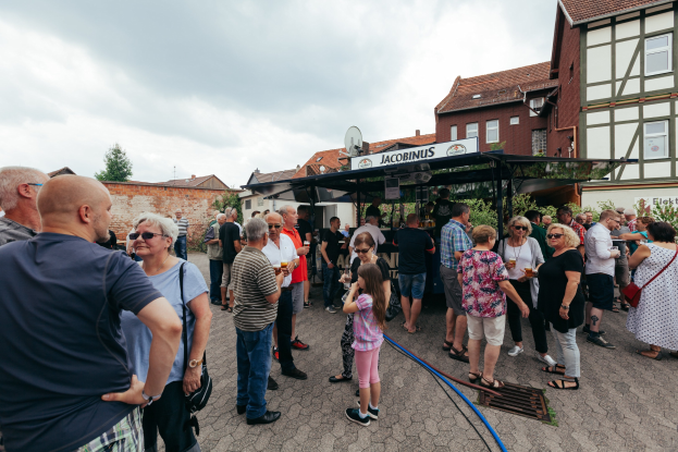 Gruppe von Menschen auf einem Outdoor-Bierfest vor einem Gebäude mit einem Schuppen, der ein Schild trägt, umgeben von Bäumen unter einem bewölkten Himmel.