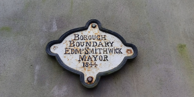 Metallschild an der Seite eines Gebäudes, das die Borough-Grenze markiert, mit "Borough Boundary Edm Smithwick Mayor" in fetten weißen Buchstaben auf schwarzem Hintergrund.