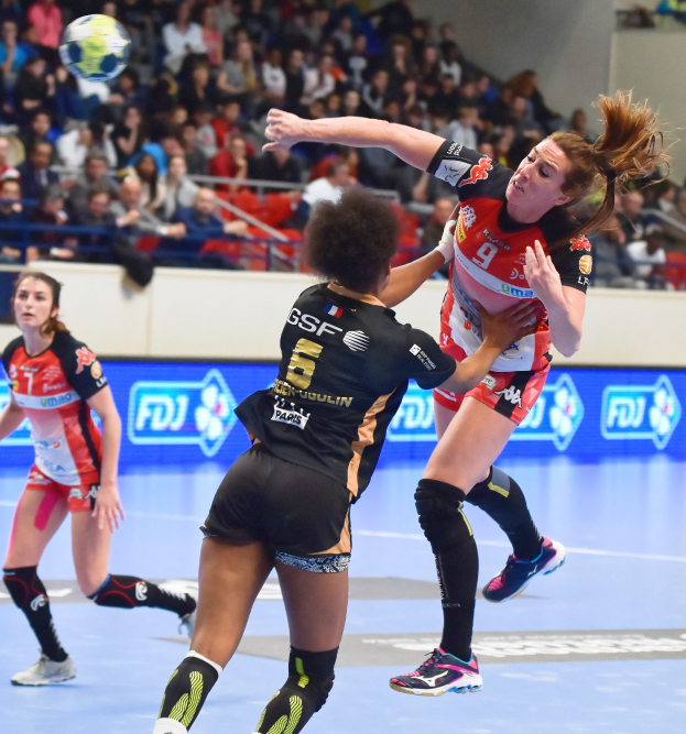 Eine Gruppe von Frauen, die auf einem Feld Handball spielen, mit einem Ball in der Luft, in einer Arena, in der Zuschauer auf Stühlen, Geländern und einer Wand sitzen.