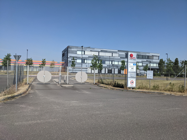 Großes Industriegebäude mit der Aufschrift "First Solar" und vielen Fenstern, umgeben von einem Metallzaun, Bäumen, Pflanzen, Gras, Schildern, Straßenmasten und einem Weg unter einem bewölktem Himmel.