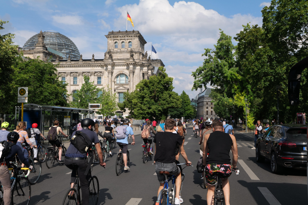 Eine Gruppe von Menschen, die auf Fahrrädern eine von Bäumen gesäumte Straße in Berlin, Deutschland, entlangfahren, mit Gebäuden und einer Bushaltestelle, unter einem bewölkten Himmel und einer Flagge, die an einem Gebäude weht.