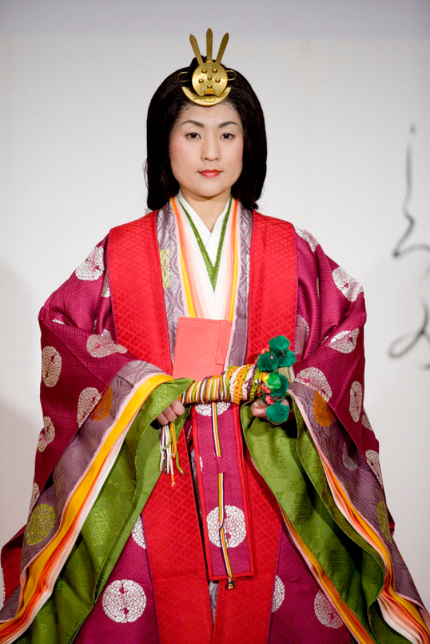Eine Frau in einem traditionellen japanischen Kimono, das ein leuchtend rotes und grünes Kleid mit aufwändigen Mustern trägt, steht selbstbewusst auf einem Laufsteg, ihr Haar ist ordentlich zu einem Knoten gebunden.