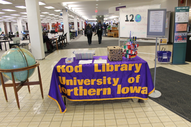 Eine Expo-Szene mit einem Tisch, der verschiedene Landesflaggen, eine Box, Papiere und eine Fahne zeigt, eine Kugel links daneben, ein Anschlagbrett rechts daneben und mehrere Tische und Stühle im Hintergrund.