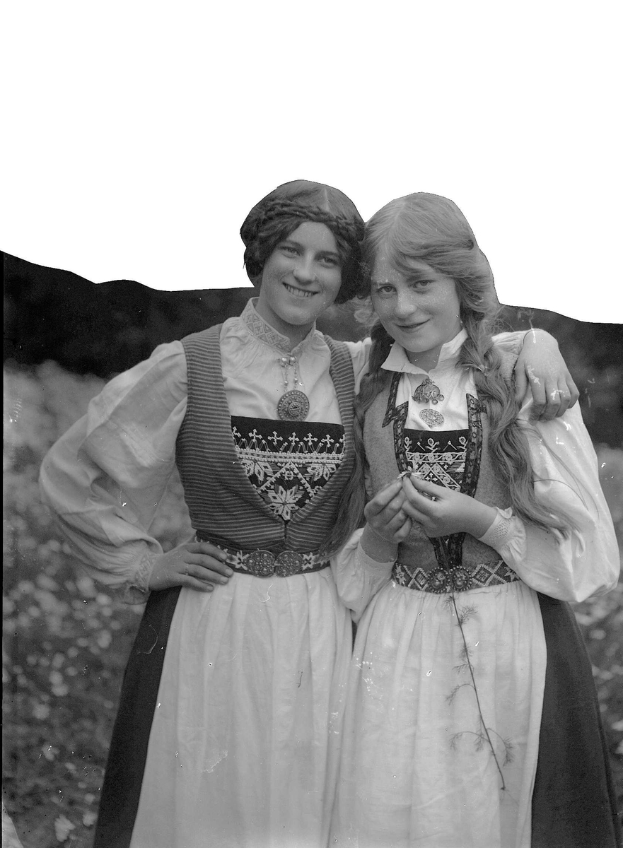 Zwei junge Frauen in traditioneller bayrischer Tracht, mit weißen und schwarzen Kleidern mit aufwendiger Stickerei und Perlen, posieren vor der Kamera und lächeln, vor einem Hintergrund aus üppigen grünen Pflanzen und einem hellen weißen Himmel.