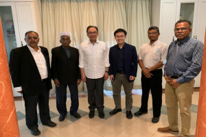 Eine Gruppe von Männern, die in einem formellen Raum mit zwei Säulen, Vorhängen und Schränken im Hintergrund zusammenstehen und über die Bildung in der muslimischen Gemeinschaft diskutieren zu scheinen.